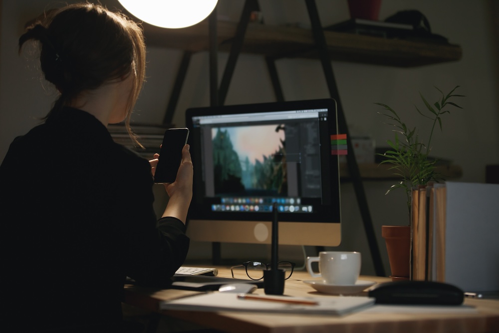Eine junge Frau bearbeitet Produktbilder am PC und hält ein Smartphone in der Hand